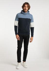 Tengerészkék és világoskék kapucnis pulóver, cowl nyakkal, kontrasztos szürke ujjak, sima textúrával, fekete, slim-fit nadrággal és fehér sportcipővel párosítva.