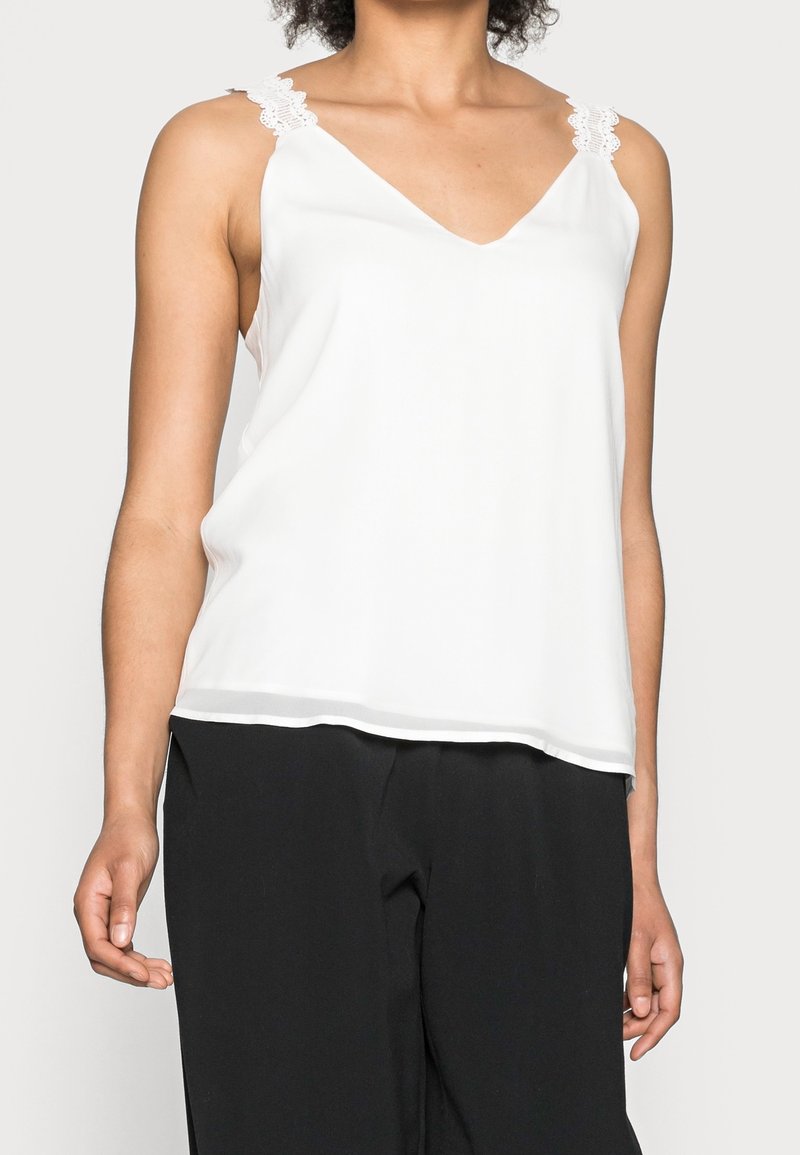 Femme portant un haut blanc sans manches avec des bretelles en dentelle et un pantalon noir, visible des épaules aux hanches sur un fond uni.