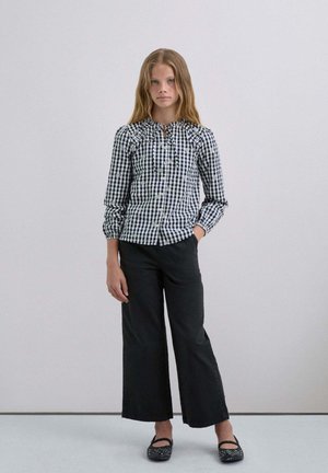 Camisa de manga larga a cuadros en blanco y negro con escote fruncido, combinada con pantalones anchos negros y zapatos planos negros con adornos.