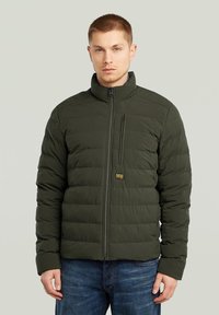 Giacca imbottita verde scuro con design a trapunta orizzontale, colletto alto e zip frontale; presenta una piccola tasca sul petto con dettaglio del logo.