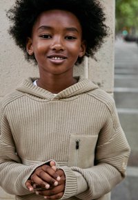 Hoodie beige en tricot avec une poche zippée à l'avant, une grande capuche et une texture côtelée, porté par un enfant avec les mains croisées devant.