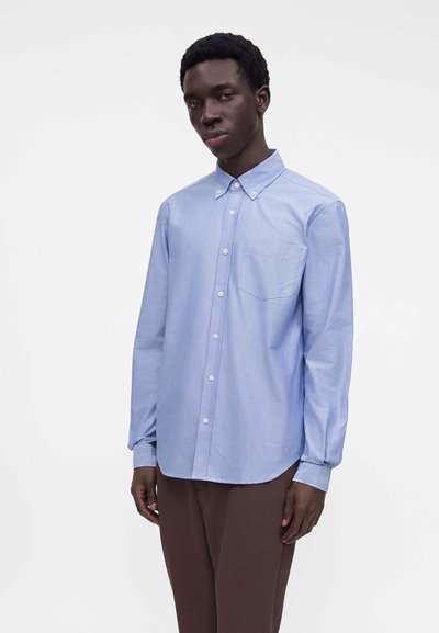 Jeune homme portant une chemise bleu clair à boutons avec une poche avant et un pantalon marron, debout devant un fond blanc uni.
