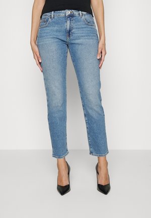 Kobieta ubrana w jasnoniebieskie jeansy o prostych nogawkach i czarne wysokie obcasy z ostrym noskiem, stojąca na tle jednolitego, jasnego tła.