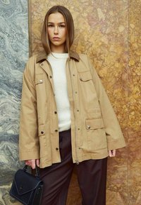 Hnedá utility bunda s kordovou golierom, s gombíkmi a zipsom, s viacerými vreckami, nosená cez smotanový sveter a hnedé nohavice. Čierna kabelka.