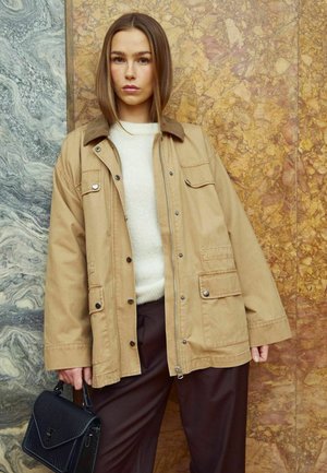 Veste utilitaire beige avec un col en velours côtelé, fermetures à boutons et à glissière, plusieurs poches, portée par-dessus un pull crème et un pantalon marron. Sac à main noir.