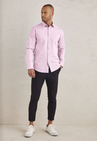 Helles rosa Langarm-Hemd mit Knöpfen, schlanker Schnitt, kombiniert mit schwarzen tapered Pants und weißen Sneakers, vor einem neutralen Hintergrund.