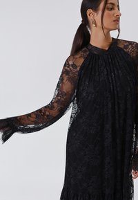 Abito di pizzo nero con lunghe maniche trasparenti, scollatura arricciata e una silhouette fluida. Presenta motivi floreali e un orlo arricciato.