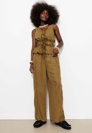 Femme aux cheveux afro naturels portant un haut sans manches vert olive et un pantalon ample avec des mocassins noirs, debout sur fond blanc.
