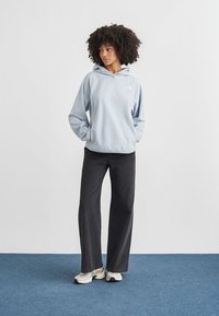 Ανοιχτό μπλε φλις φούτερ με κουκούλα, εμπρόσθια τσέπη και λογότυπο. Συνδυασμένο με μαύρο παντελόνι φαρδύ στη μπάκα και λευκά αθλητικά παπούτσια. Απλό φόντο.