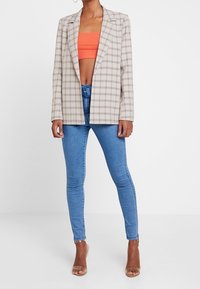 Blazer à carreaux beige avec épaules structurées, devant ouvert et manches longues. Associé à un jean bleu ajusté et un crop top orange.