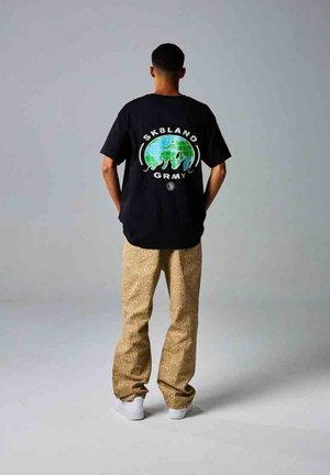 Camiseta negra con un gráfico colorido de un globo y el texto "SKBLAND GRMY"; combinada con pantalones beige con patrón y zapatillas blancas.