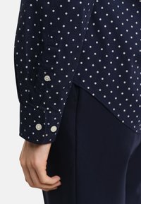 Navy shirt with white polka dots, featuring buttoned cuffs and a curved hem. Fabric appears smooth, with visible stitching detail.