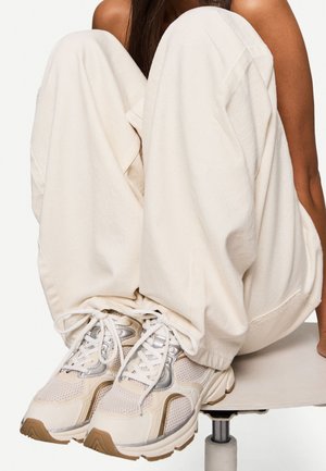 Persoon zittend met opgetrokken knieën, draagt losse witte broek en beige-zilveren veterschoenen, gezeten op een kruk of stoel.