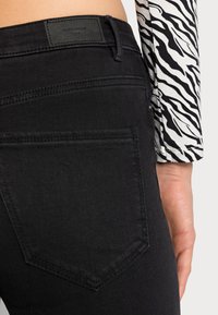 Jeans de mezclilla negra con una textura suave, que cuentan con bolsillos traseros y una etiqueta de cuero. Se puede ver una blusa de manga larga rayada de blanco y negro con estampado de cebra.