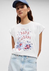 Λευκό βαμβακερό T-shirt με κοντά μανίκια-καπέλο και χρωματιστό λουλουδάτο και φραουλένιο γραφικό σχέδιο· συνδυασμένο με ανοιχτό μπλε τζιν σορτς.