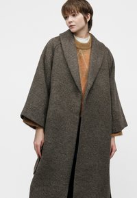 Young person wearing a loose, long dark gray patterned coat over a brown sweater, standing against a plain light background, looking right.