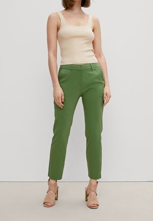 Kvinde iført beige ribbet tanktop, grøn skræddersyet ankel-længde bukser og nudefarvede højhælede sandaler, stående mod en ensfarvet baggrund.