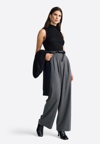 Zwarte mouwloze top, grijze hoog wassende geplooide broek, zwarte riem en zwarte enkellaarsjes. Model houdt een zwarte trui vast.