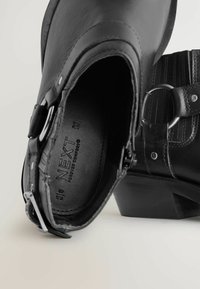 Botas de tobillo de cuero negro con un hebilla lateral, detalles texturizados y un tacón bajo apilado. Marca interior visible.