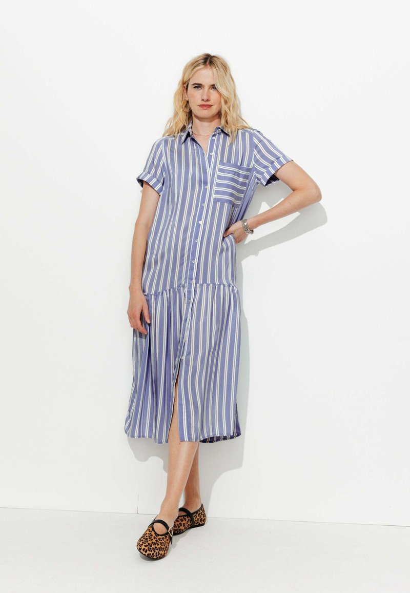 Robe rayée en tissu bleu clair et blanc, avec un design à boutonnière, une poche, des manches courtes et une jupe à volants. Associée à des chaussures à imprimé léopard.