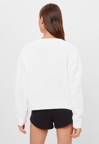 En kvinna med långt brunt hår bär en vit stickad tröja och svarta shorts, ståendes vänd bort mot en enfärgad ljus bakgrund.