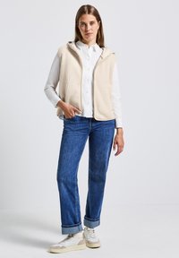 Gilet en polaire beige avec capuche, fermeture à glissière, associé à une chemise blanche et un jean bleu. Baskets blanches avec des accents beiges. Design minimaliste.
