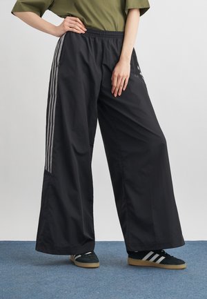 Personne portant un pantalon noir à jambes larges avec des bandes grises sur les côtés, des baskets noires avec des bandes blanches, debout sur un tapis bleu.