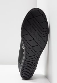 Černá podrážka tenisky s dezénem a embosovaným nápisem "Calvin Klein Jeans", postavená vzpřímeně proti bílé rohové zdi.