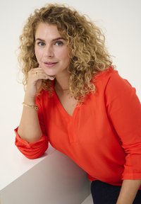 Frau mit lockigem Haar, die ihr Kinn auf ihre Hand stützt, trägt eine leuchtend orangefarbene Bluse mit V-Ausschnitt aus strukturiertem Stoff und sitzt vor einem neutralen Hintergrund.