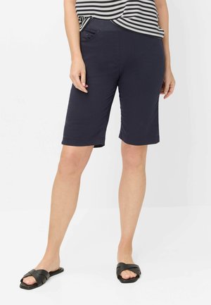 Kvinde iført marineblå knælange shorts, en sort-hvid stribet top og sorte slip-on sandaler, stående mod en hvid baggrund.