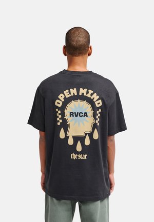 Uomo con capelli corti che indossa una maglietta nera con una grafica beige di una testa e il testo "OPEN MIND RVCA the star" sul retro.