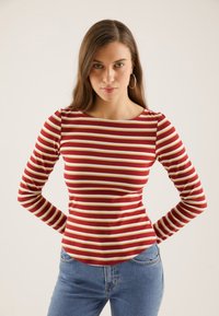 Rød langermet topp med kremfargede og gull horisontale striper, med rund hals og tettsittende design, kombinert med blå denimjeans.