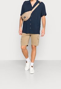 Camicia button-up a maniche corte blu navy abbinata a pantaloni cargo beige. Sneakers bianche e borsa a tracolla beige. Texture lisce, vestibilità casual.