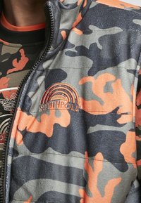 Gros plan d'une personne portant une veste camouflage orange, grise et noire avec une fermeture éclair noire et un logo "Southpole" brodé avec un design arc-en-ciel.