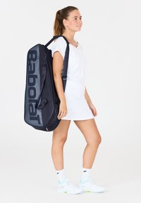 Schwarze Tennistasche mit großem grauen "Babolat"-Logo, gepolsterten Griffen und verstellbarem Schultergurt, getragen von einer Person in einem weißen Outfit.
