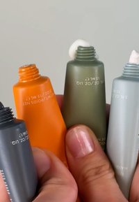 Fire squeeze-tuber i oransje, grå, olivengrønn og lysegrå, hver merket med 15 ml, holdt mellom fingrene med naturlige negler.