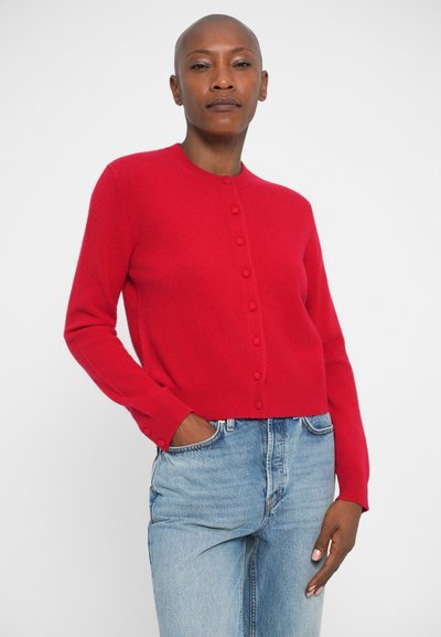 Cardigan roșu tricotat cu guler rotund, închidere cu nasturi în față și mâneci lungi. Asortat cu blugi din denim de culoare albastră deschisă.