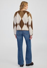 Braun-weißer Argyle-Muster-Wollpullover, Rundhalsausschnitt, flauschige Textur, kombiniert mit hochgeschnittenen blauen Schlagjeans und dunklem Schuhwerk.