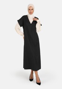 Black modest dress with a V-neck and short sleeves over a cream blouse, featuring smooth fabric, relaxed fit, and ankle-length design.