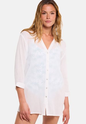 Mujer con cabello hasta los hombros que lleva una camisa blanca transparente abotonada con las mangas remangadas sobre un fondo liso.
