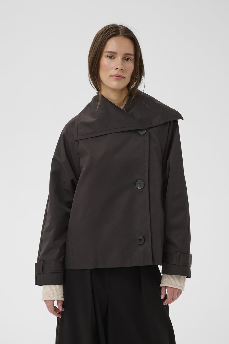 Donna che indossa una giacca scura oversize con ampio colletto e grandi bottoni, in piedi contro uno sfondo chiaro e semplice.