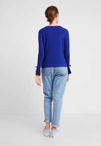 Blaues Langarmoberteil mit gebundenen Manschetten, kombiniert mit hellen Jeans. Das Oberteil hat eine lockere Passform und einen leicht abgerundeten Ausschnitt.