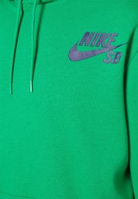 Zelená mikina s kapucí, vyrobená z jemného materiálu; na přední straně má šedé logo Nike SB. Obsahuje nastavitelné šňůrky s kovovými konci.