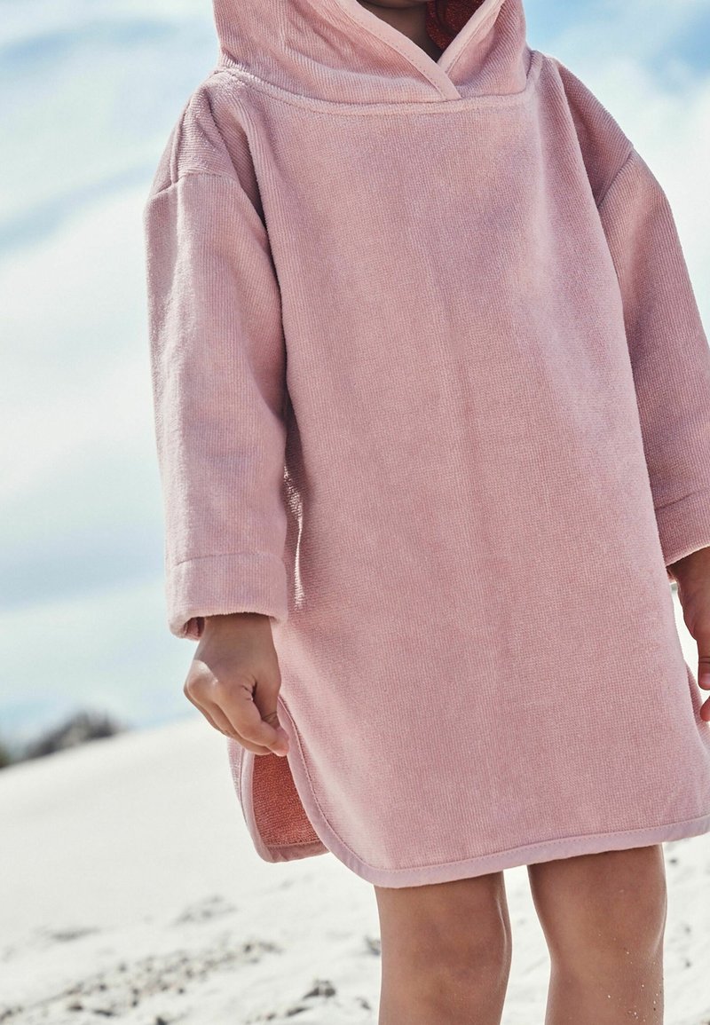 Enfant debout sur une plage sableuse portant une robe serviette à capuche rose ample avec manches longues et fentes latérales.