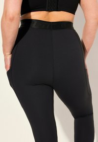 Leggings neri con vita alta, dotati di tasche laterali e un tessuto morbido e elasticizzato che adatta perfettamente le gambe. Le cuciture sono chiaramente visibili.
