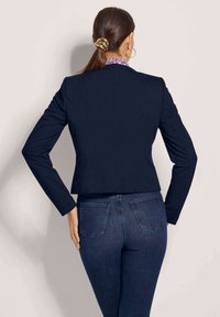 Marineblaues, kurzes Jacket mit strukturierten Nähten und langen Ärmeln. Über dunklem Denim getragen, mit einem gemusterten Kragen und subtilen Details.