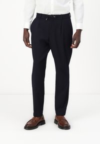 Pantaloni blu tailored con vita regolabile, frontale plissettato e vestibilità slim. Indossati con mocassini marroni e una camicia bianca con button-up.