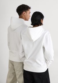 Bijele dukserice s kapuljačom, opuštenog kroja, s rebrastim manžetama i rubovima. Jedna dukserica je uparena sa bež hlačama, dok je druga uparena s crnim hlačama.