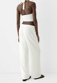Bílé texturované kalhoty širokého střihu s elastickým pasem, doplněné bílým halter topem se zavazováním na zádech. Viditelné černé sandály.