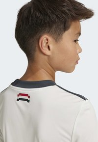 Camiseta atlética branca com gola escura, apresentando um pequeno logótipo vermelho, branco e azul na parte de trás. Tecido suave, mangas curtas, design simples.